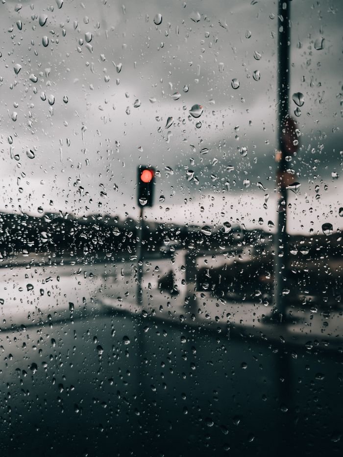 our-story Close-up of raindrops on a window with a red traffic light in the background, capturing a rainy day mood.